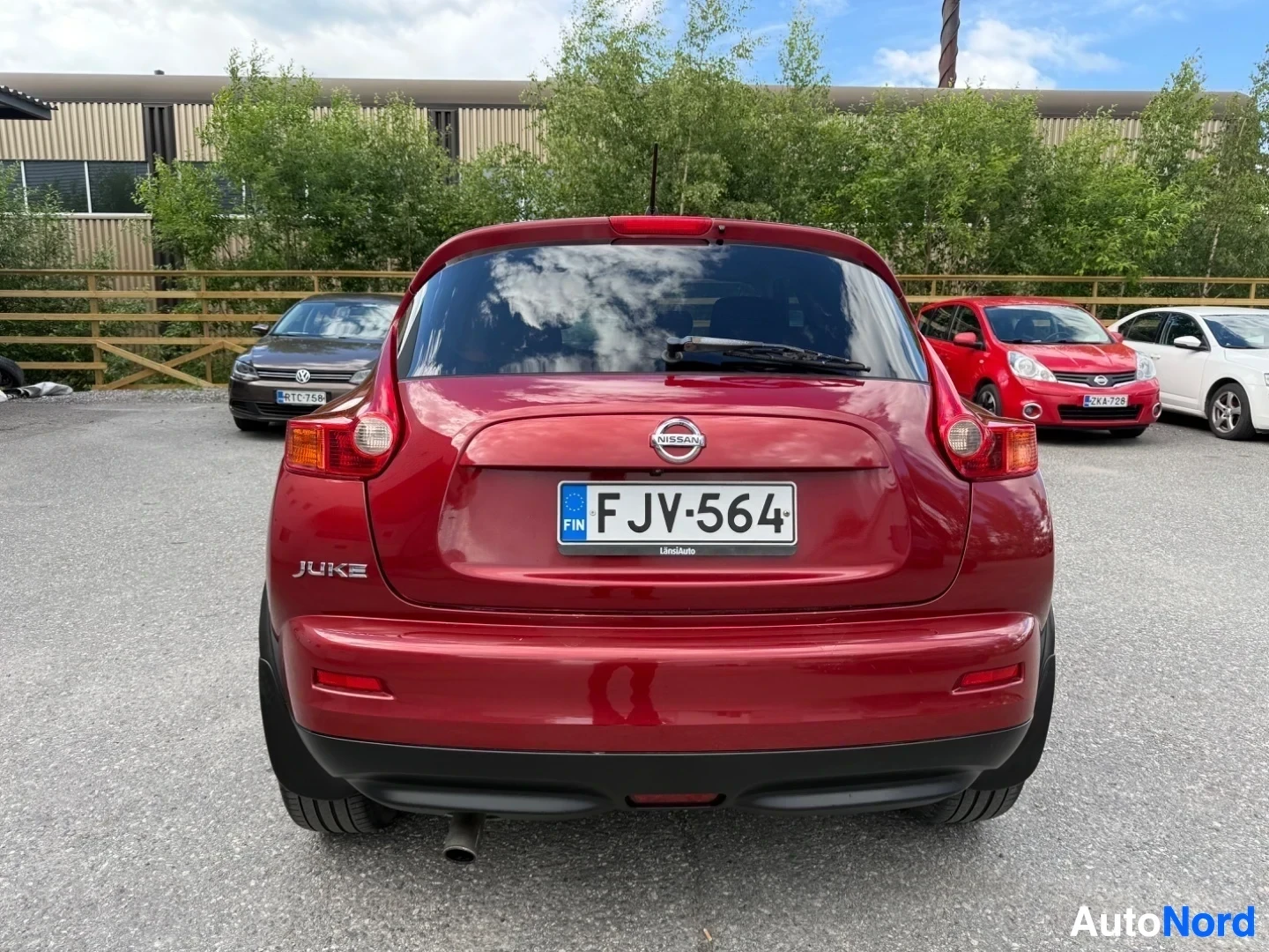Takaa: Punainen Nissan Juke takavasemmalta kuvattuna. Näkyy rekisterikilpi FJV564 ja merkinnät "JUKE" sivulla. Taustalla muita autoja ja rakennuksia.