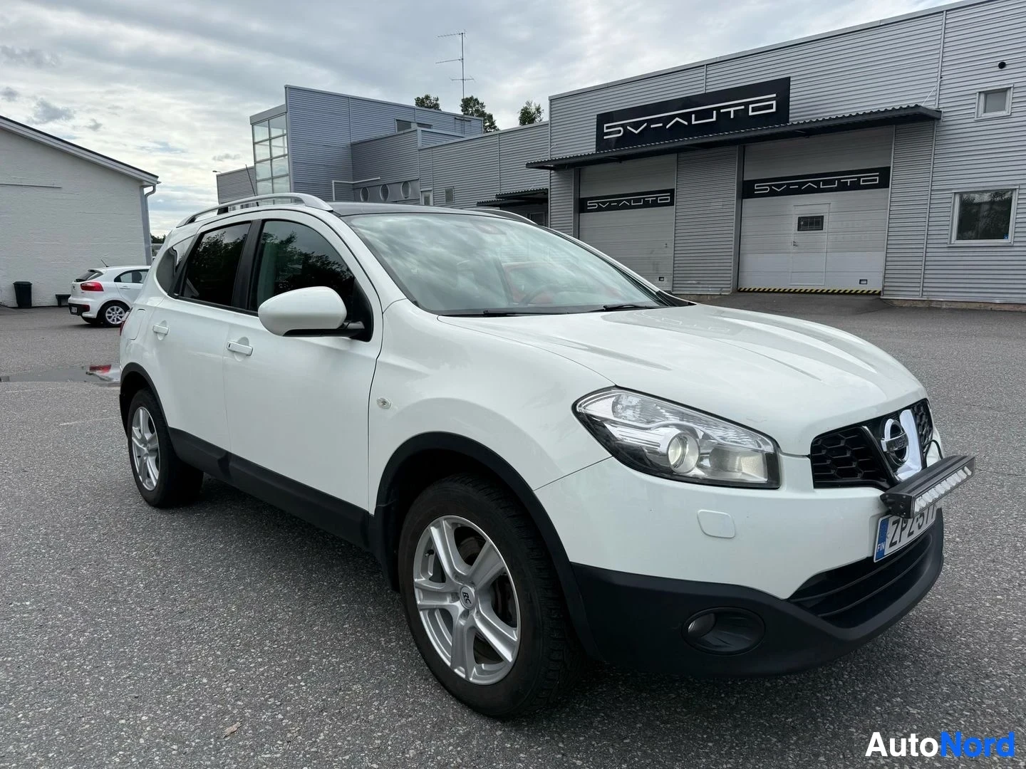Etuviisto, nokka oikealle: Valkoinen Nissan Qashqai etupuoli kolmen neljäsosan kulmasta, oikea puoli edessä, teollisuusrakennuksen edessä pysäköity