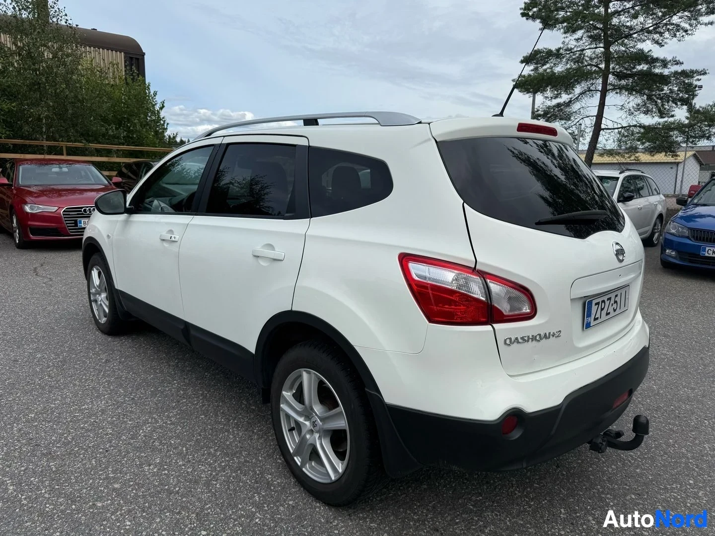 Sivulta: Valkoinen Nissan Qashqai+2 -auto oikealta sivulta katsottuna parkkipaikalla. Näkyvät avoin kuljettajan ovi, punaiset takavalo, hopeasärmät ja FIN rekist