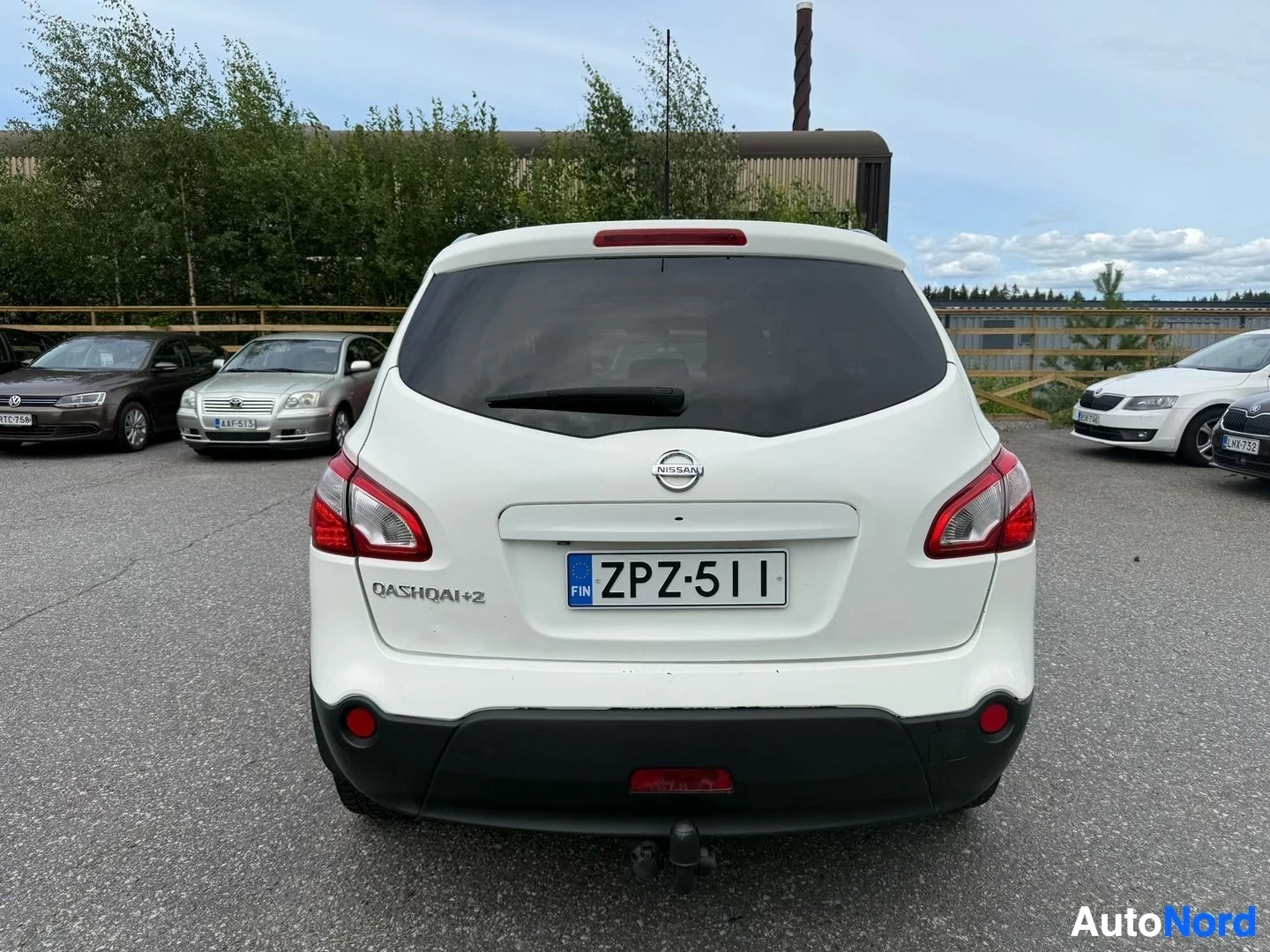 Takaa: Valkoinen Nissan Qashqai+2 -auto takaa katsottuna parkkipaikalla. Rekisterikilvessä FIN. Punaisen ja valkoisen takavalojen ja mustan alapalokin näkyvät