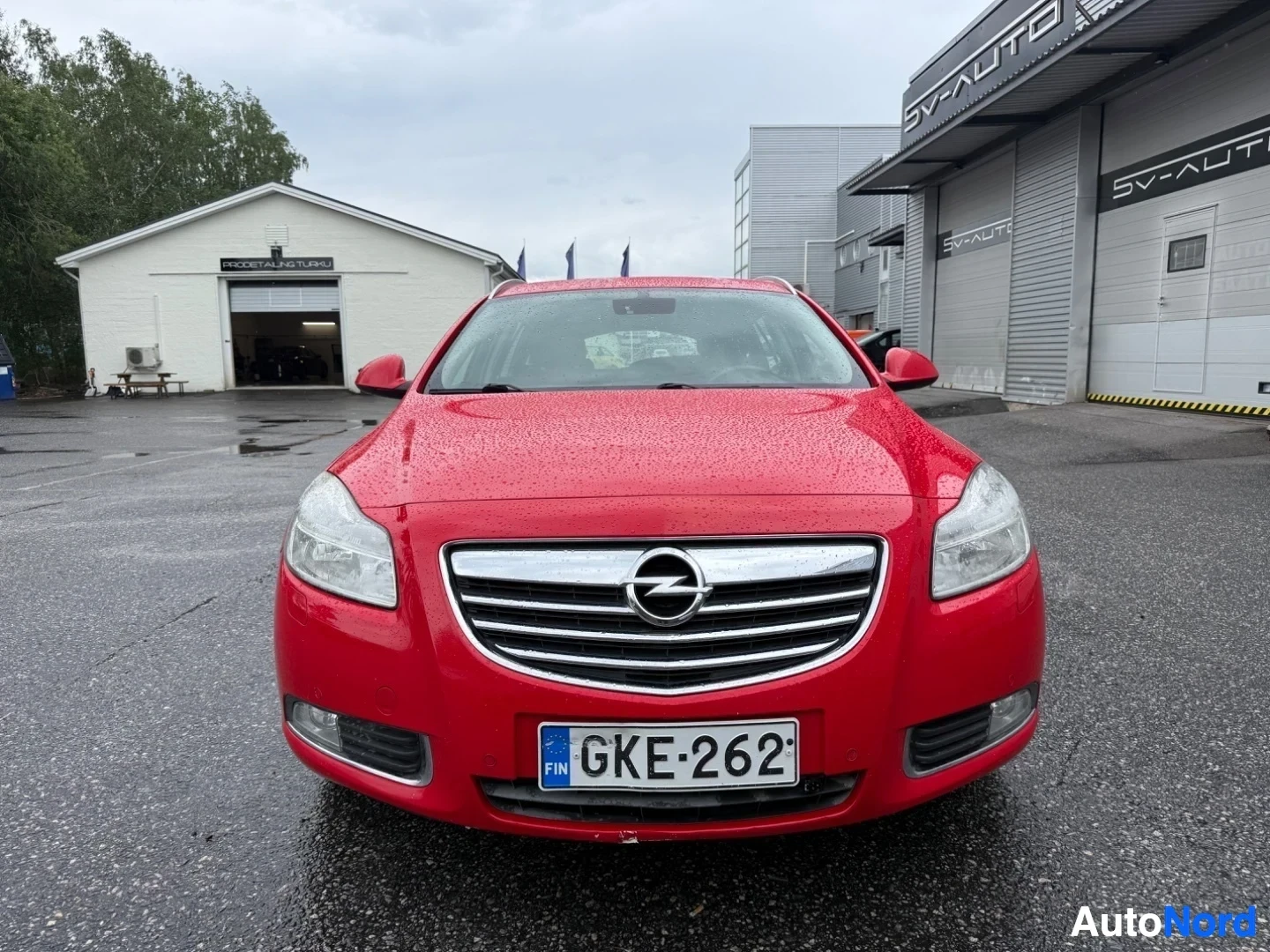 Edestä: Punainen Opel Insignia etunäkymä, mustalla grillillä ja valkoisen rekisteröintikilven kanssa, pysäköity asfalttialueella rakennusten edessä