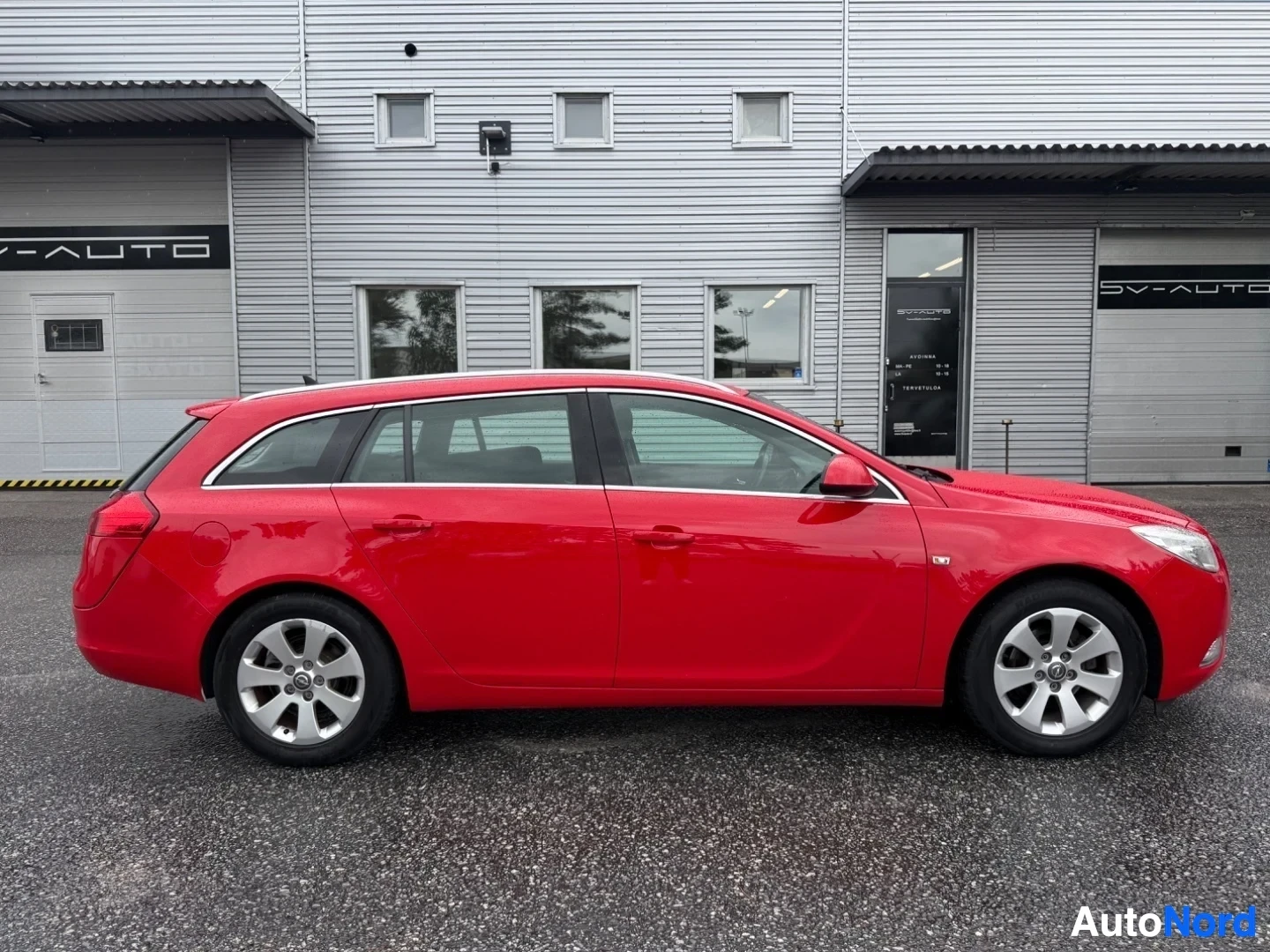 Sivulta: Punainen Opel Insignia Sports Tourer profiilissaan oikealle, täysi sivunäkymä myyntiliikkeen edessä.