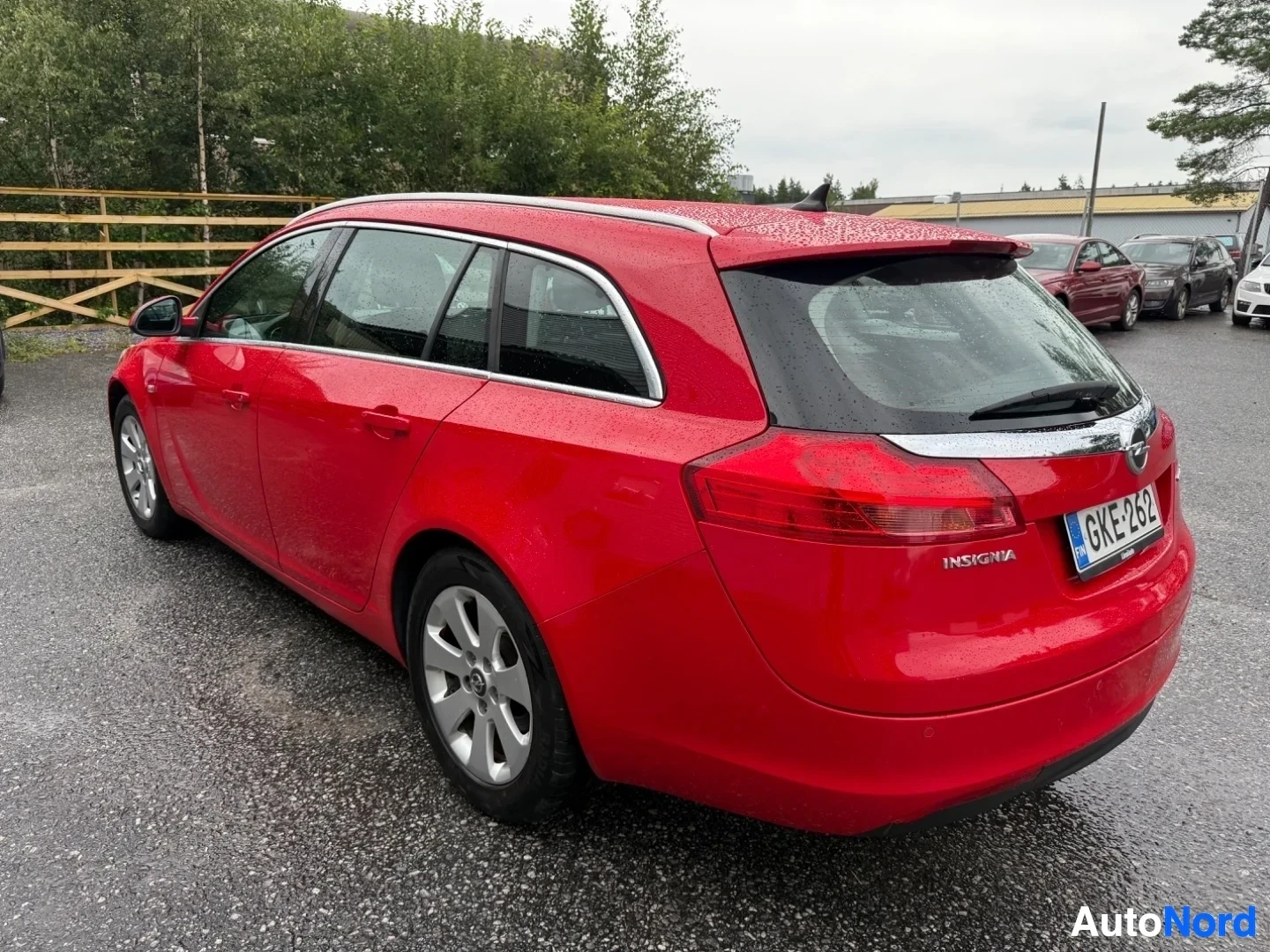 Sivulta: Punainen Opel Insignia station wagon sivusta, rekisteröintinumero näkyvissä, valkoinen rengas-levy näkyy, pysäköity asfalttialueella