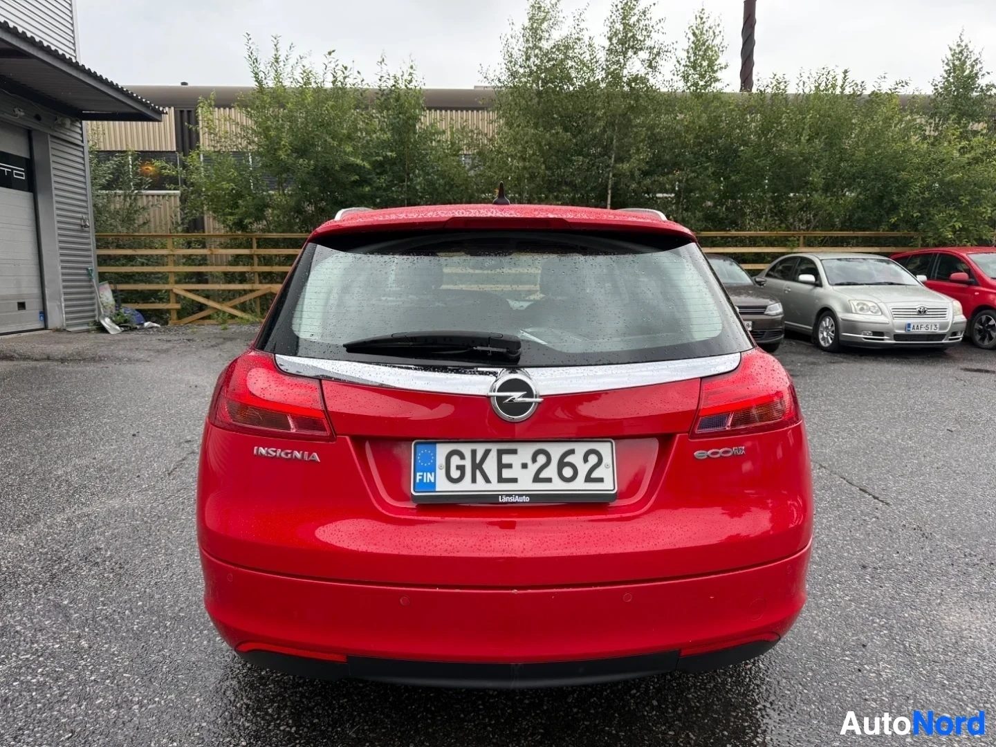 Takaa: Punainen Opel Insignia station wagon ajoneuvon takaosa, rekisteröintinumero näkyvissä, pysäköity asfalttialueella pihattujen rakennusten edessä