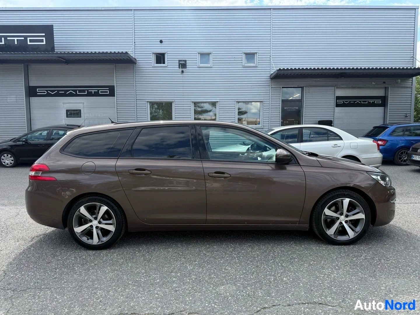 Sivulta: Ruskea Peugeot 308 SW farmari sivusta kuvattu, seisoo jäljellä olevien autojen edessä myyntilotolla.