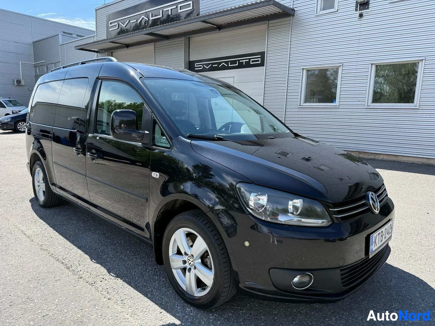 Etuviisto, nokka oikealle: Musta Volkswagen Caddy -pakettiauto etukulman näkymässä, kunnostamon edessä