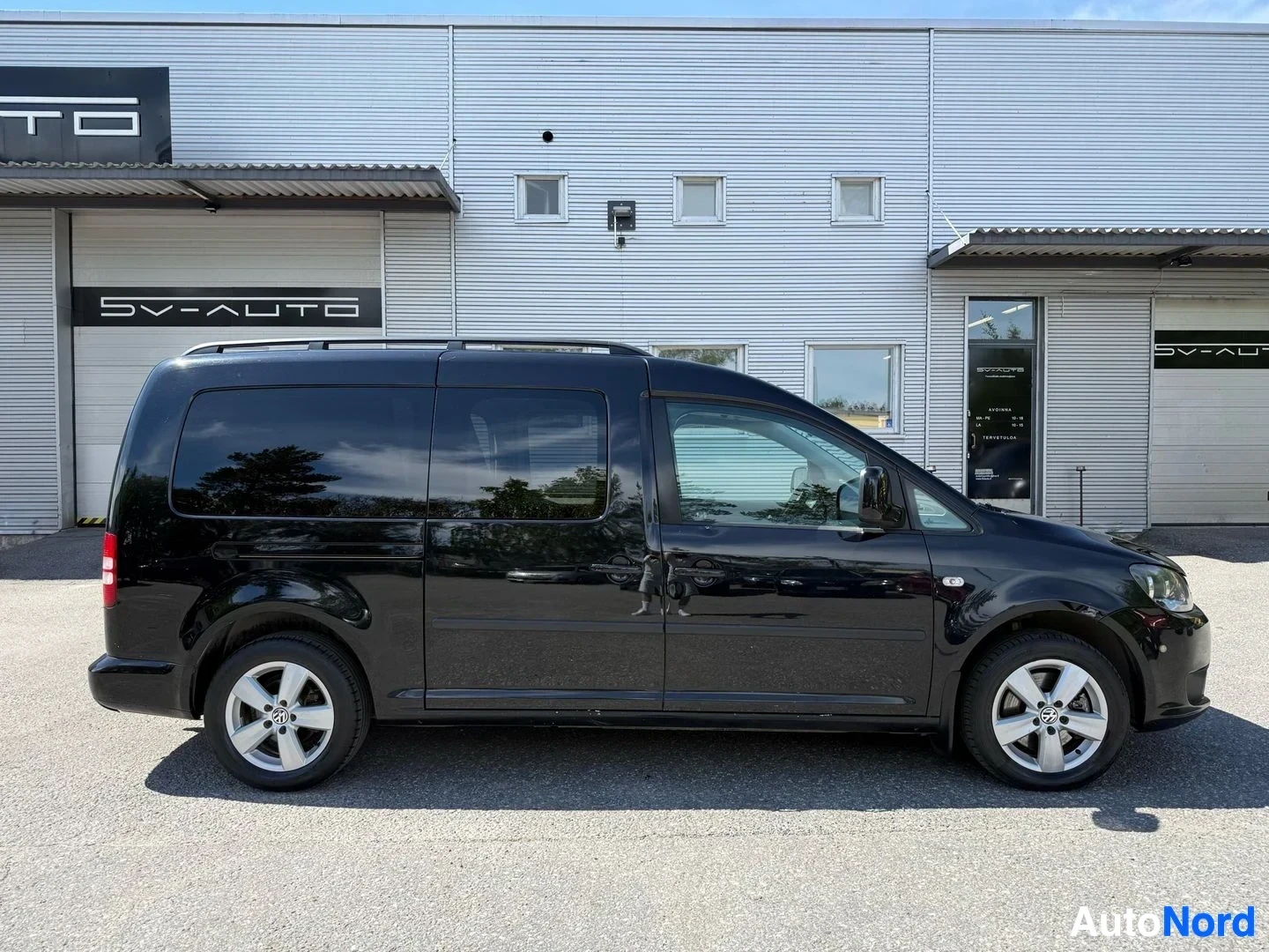 Sivulta: Musta Volkswagen Caddy -pakettiauto sivuprofiilissa, kunnostamon seinän edessä