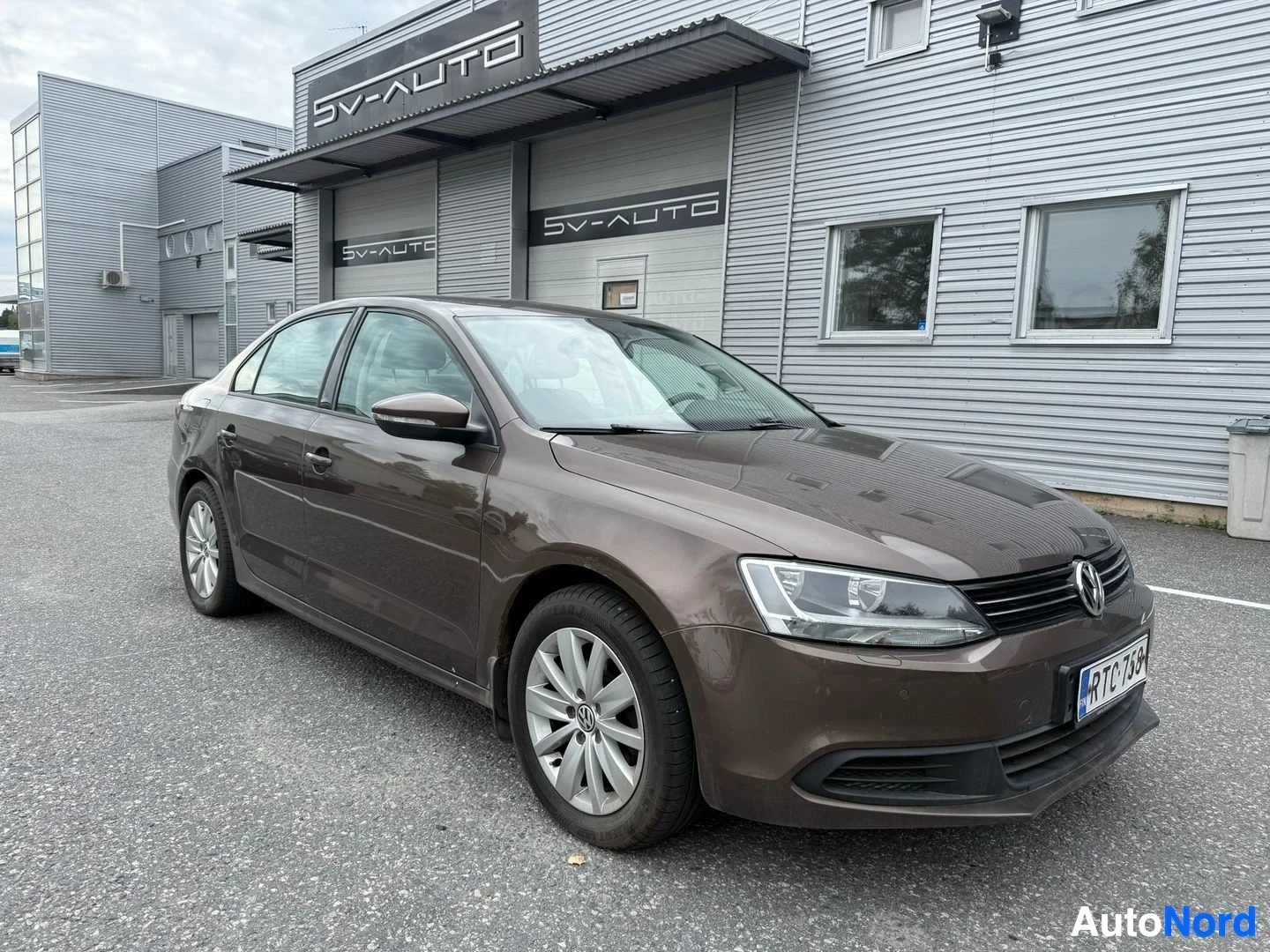 Etuviisto, nokka oikealle: Ruskea Volkswagen Jetta, etupuoli oikealle katsoen, SV-Auto myyntitallin edessä.