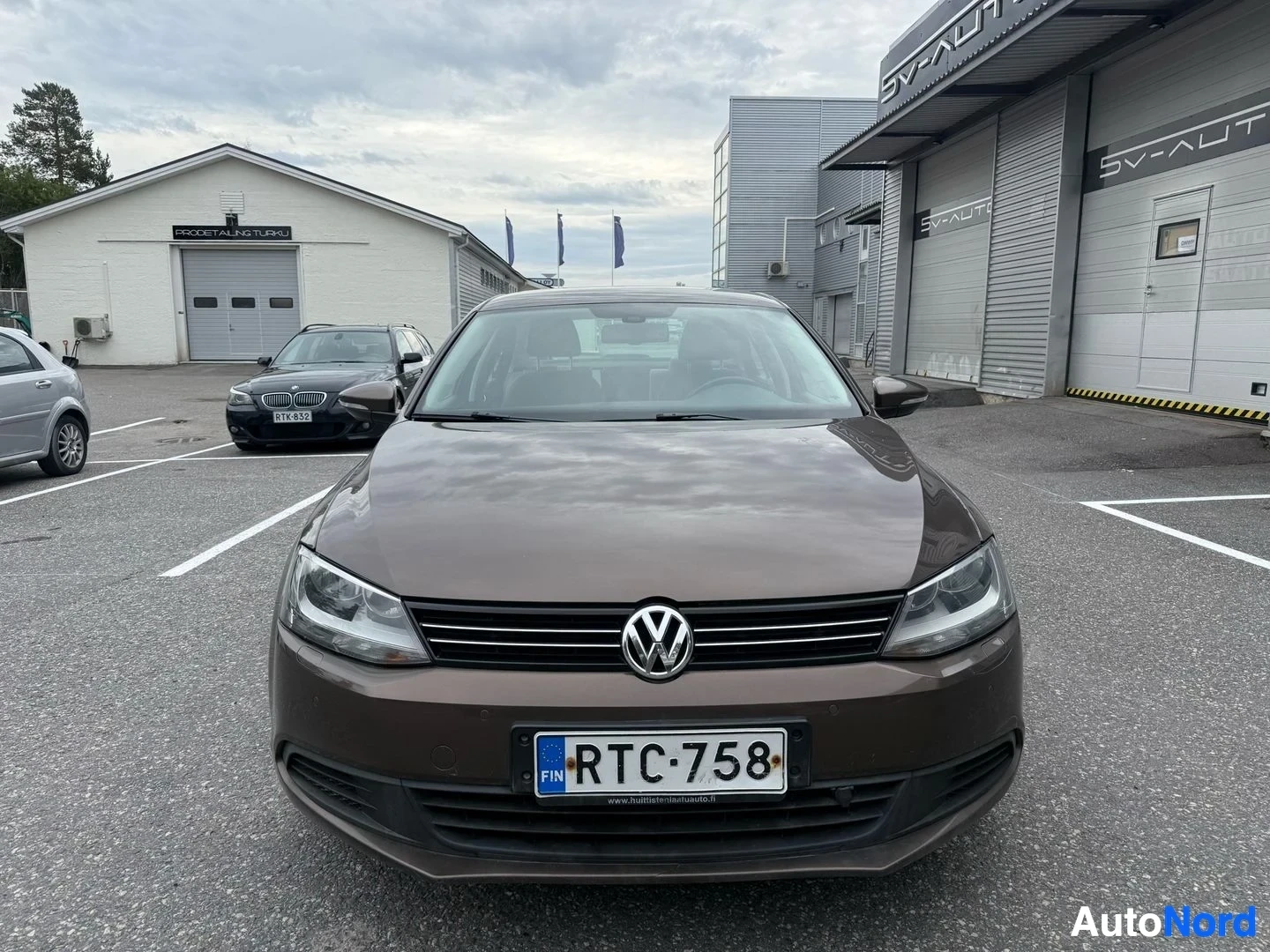 Edestä: Volkswagen Jetta edestä katsottu, ruskea sedan pysäköitynä parkkipaikalle, rekisterikilvessä, hopeisiset ajovalot näkyvissä