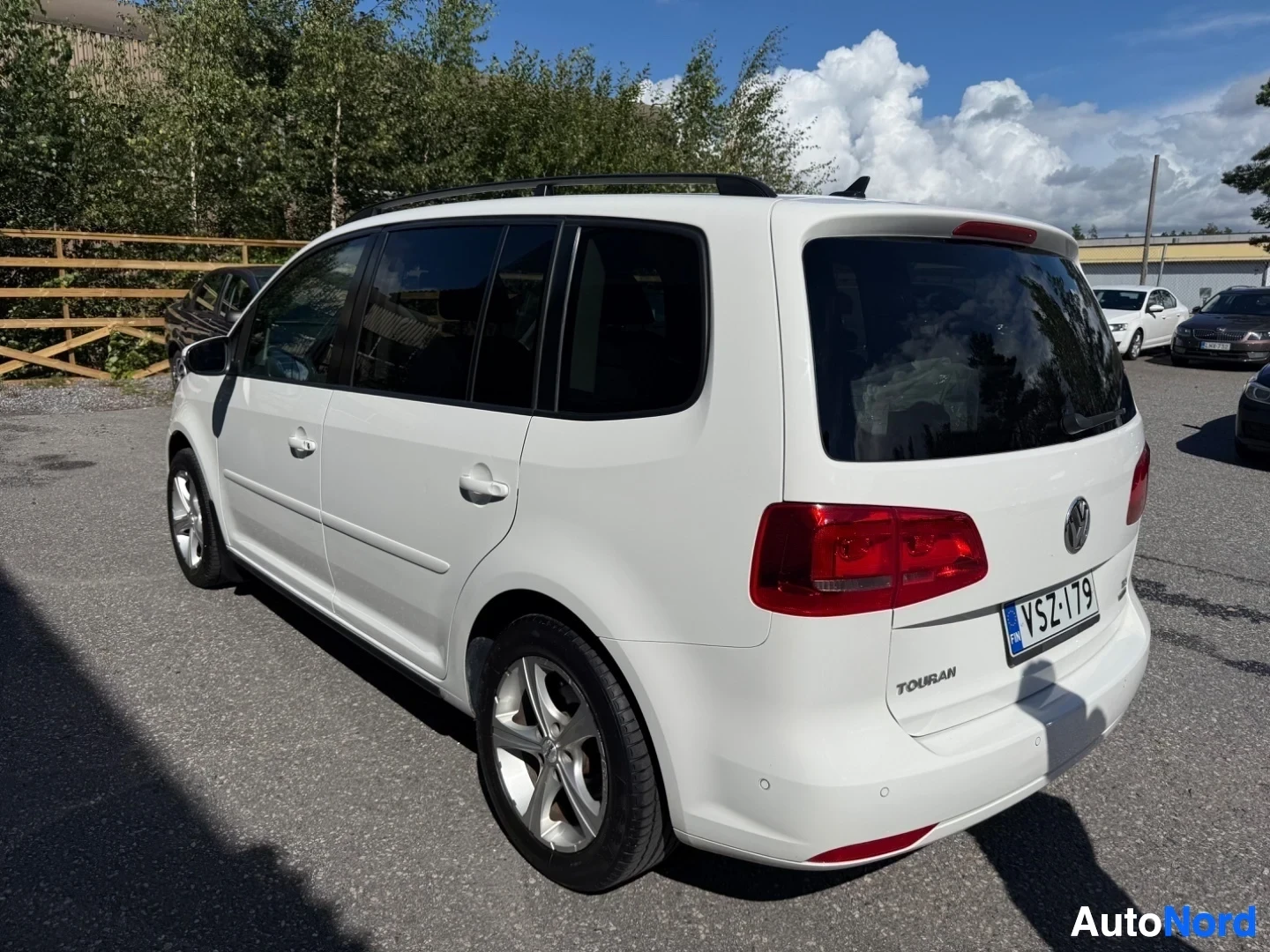 Takaviisto: Valkoinen Volkswagen Touran kolme neljäsosaa takaa oikealta, punainen taka-valo, rekisterinumero näkyy