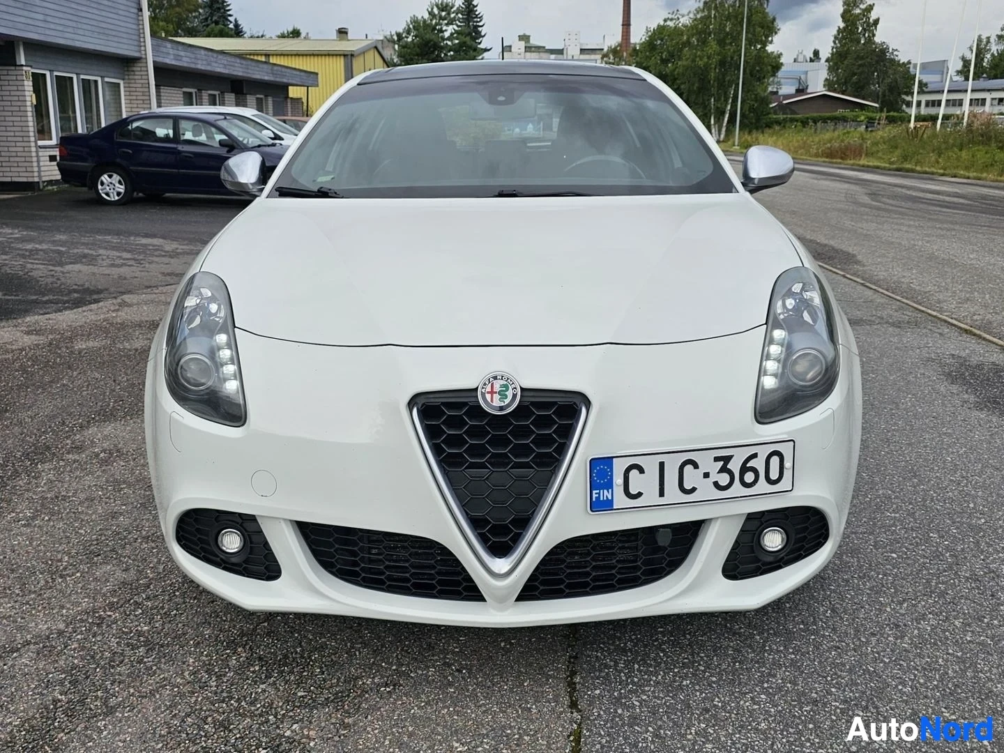 Edestä: Valkoinen Alfa Romeo Giulietta etupuolelta, kuvattuna suoraan edesta pysäköintialueella. Näkyy Suomalainen rekisterikilpi