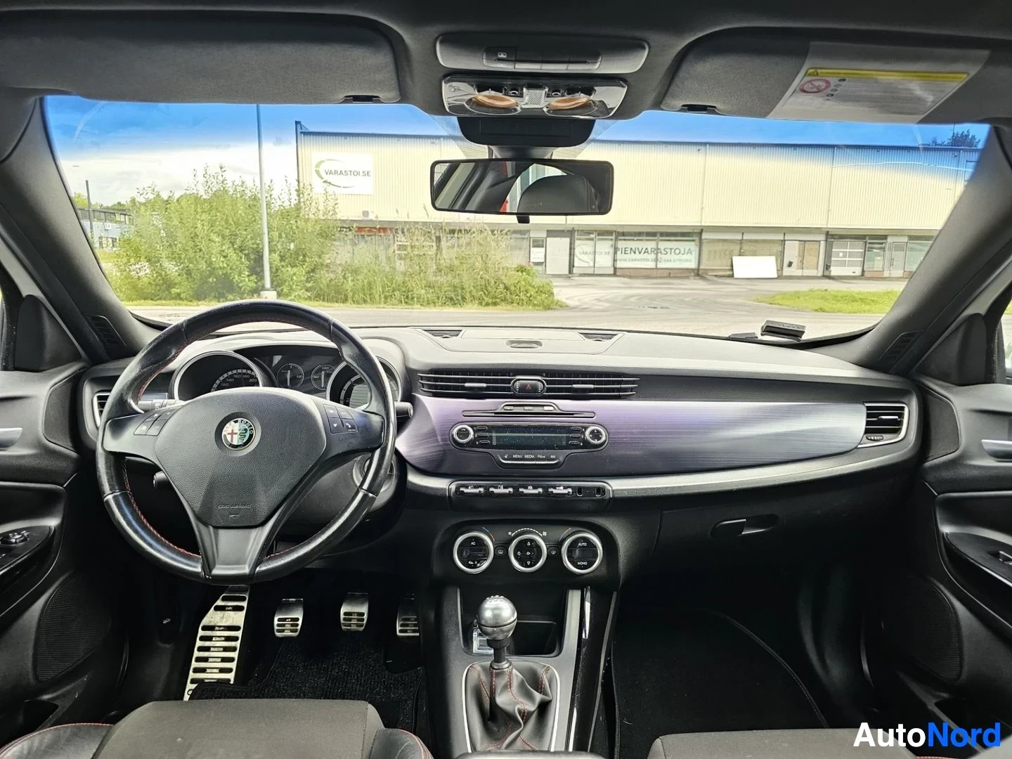 Ohjaamo: Ohjaamossa Alfa Romeo-ohjauspyörä, paljastettu ilmastointi-nappulat, radio näytöllä punaisella tekstillä, musta sisustus, panoraamamainen näkymä edestä