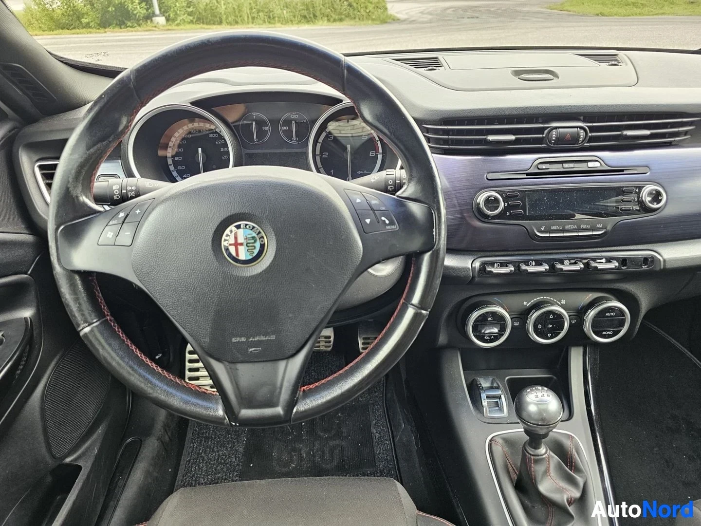 Yksityiskohta: Ohjauspyörä lähikuvassa punaisella vaaralla, mittaristo taustalla, Alfa Romeo-logo pyörän keskuksessa, ilmastointi-nappulat oikealla