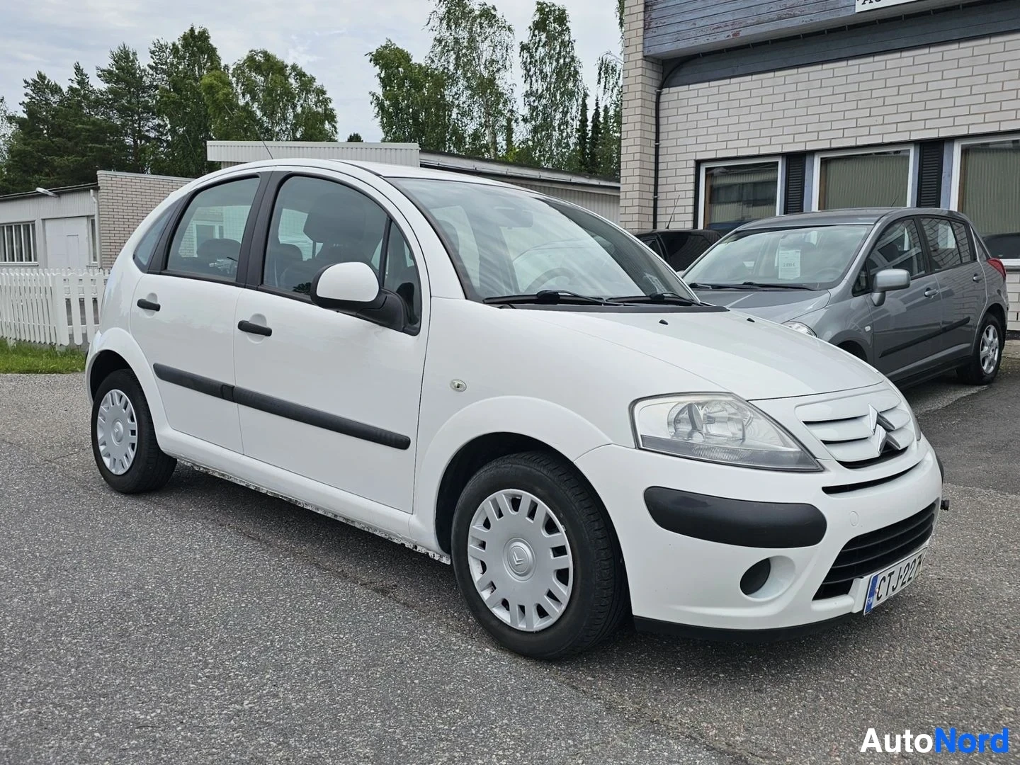 Etuviisto, nokka vasemmalle: Valkoinen Citroën-pieni viistoperä paikoitusalueella, oikeasta kulmasta kuvattuna. Auto näkyy kokonaan, valkoinen väri, mustat raidat sivuilla.