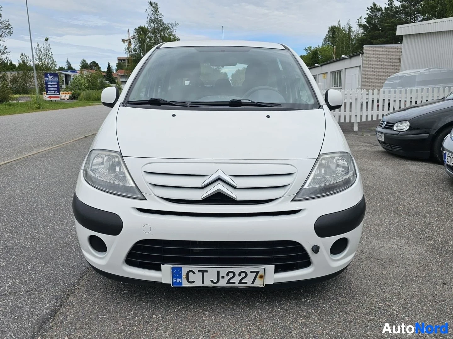 Edestä: Valkoinen Citroën-pieni viistoperä kuvattuna suoraan edestä paikoitusalueella. Auton koko etuosa näkyy selvästi.