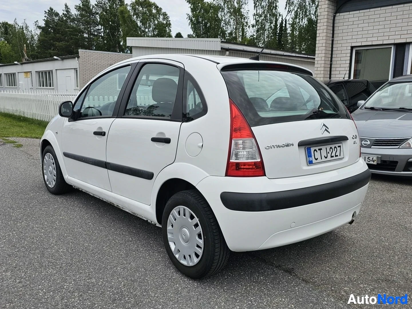 Takaviisto: Valkoinen Citroën C3, takavasemmalta näkymä, pysäköitynä pihalla puiden lähellä