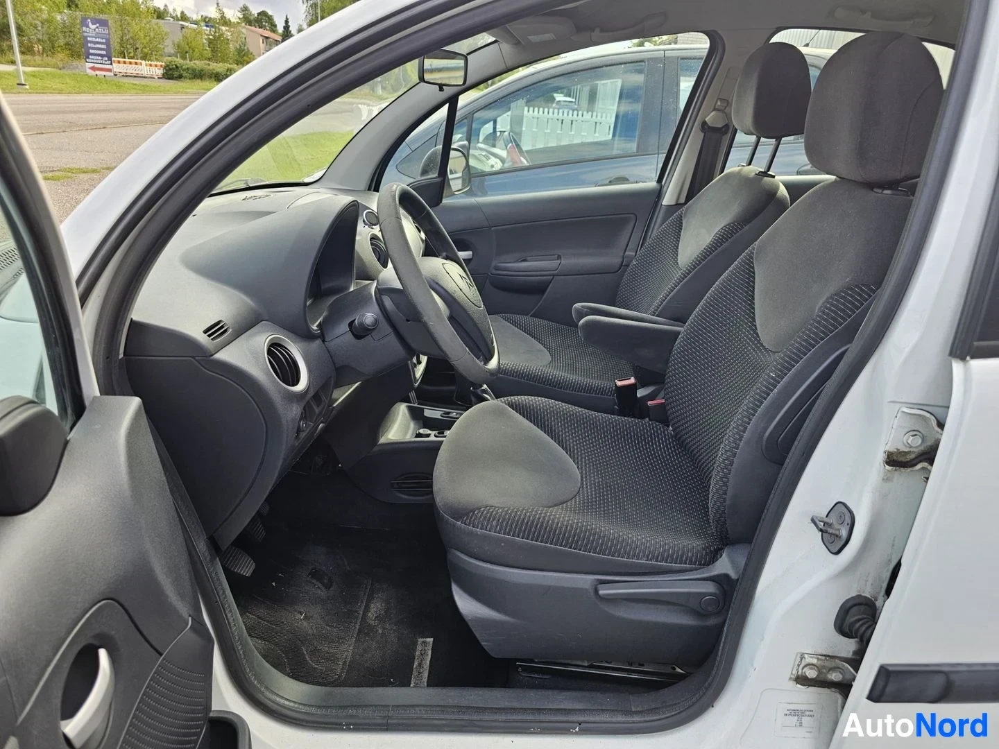 Ohjaamo: Valkoisen Citroën C3:n sisusta, ovenaukosta otettu kuva, näkyy musta ohjauspyörä, mustat kangaspäällysteisen istuinten, elektroninen parkkiristikko ja mustalla