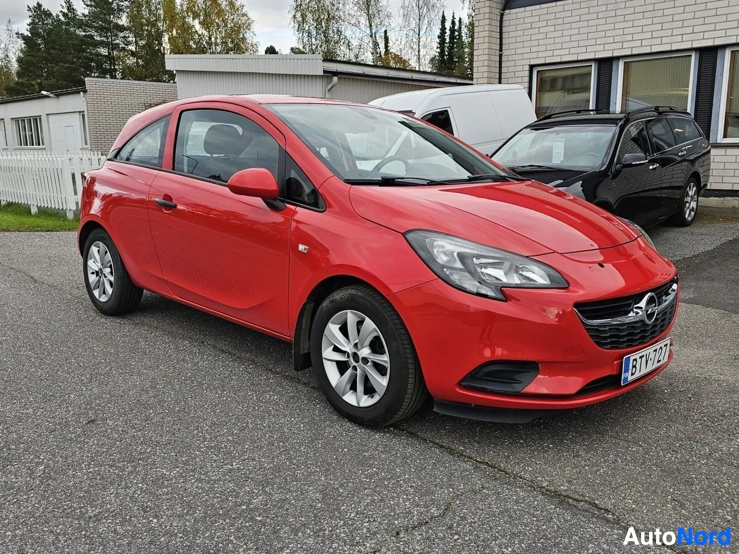 Etuviisto, nokka vasemmalle: Punainen Opel Corsa kolmen hengen pakettiauto, etukolmikulmaviistosta vasemmalta nähtynä, valkoilla vanteeilla, pysäköitynä tallirakennuksen edessä.