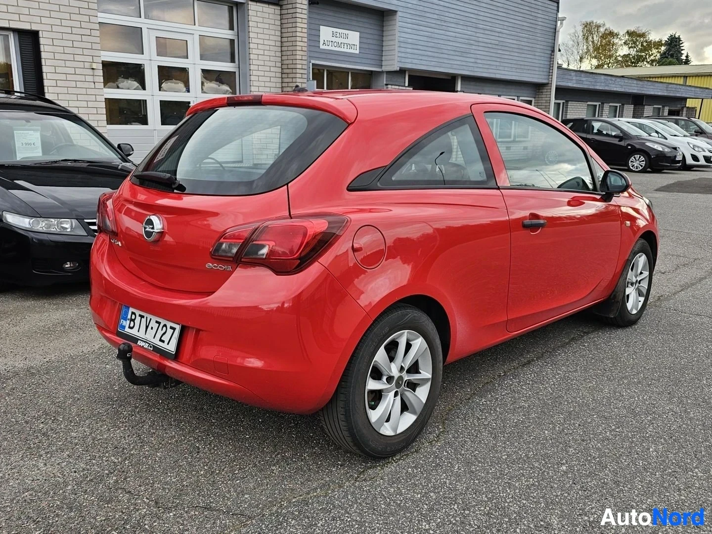 Takaviisto: Punainen Opel Corsa kolmen hengen pakettiauto, takaviistosta vasemmalta nähtynä, valkoilla vanteeilla, pysäköitynä liikepaikalla.
