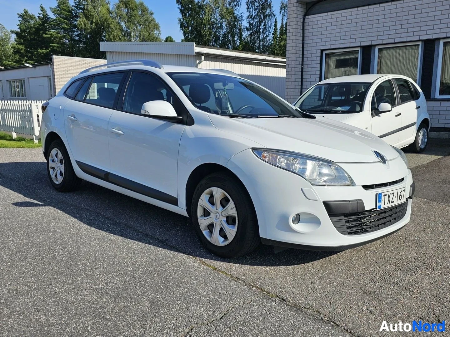 Etuviisto, nokka vasemmalle: Valkoinen Renault Megane Sport Tourer -farmari sivusta kuvattuna pysäköitynä asfaltille. Rekisterikilvessä