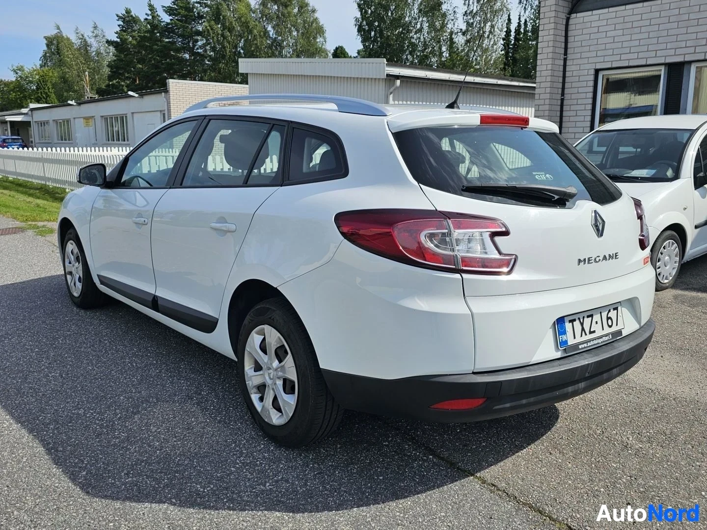Takaviisto: Valkoinen Renault Megane farmari, takaa vasemmalta, parkkipaikalla nurmikon vieressä, vasen ovi auki