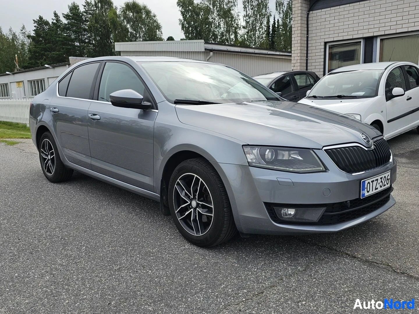 Etuviisto, nokka vasemmalle: Hopeanvärinen Skoda-merkkinen sedan, oikealle kääntynyt kolmen neljänneksen kuva, näkyy koko ajoneuvo sivulla, metalliset vanteet, tummennettuja ikkuno