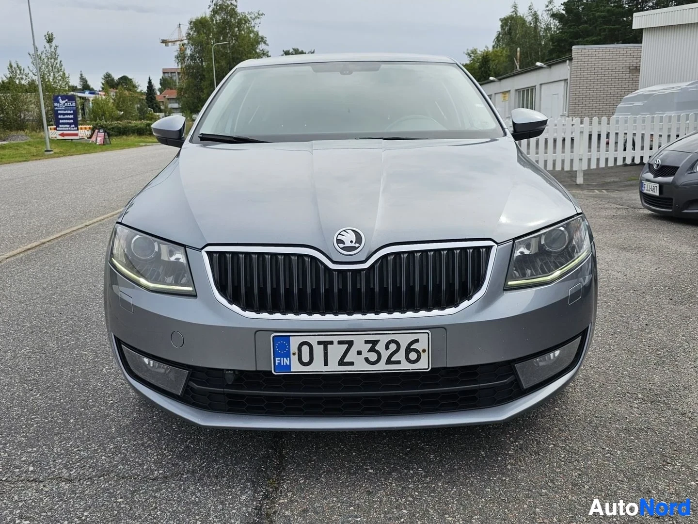 Edestä: Hopeanvärinen Skoda-sedan etuosasta suoraan kuvattuna, mustavalkoinen pystysuuntainen säleikkö etuosassa, LED-päivänvalot näkyvät, rekisterikilpi näkyvissä.