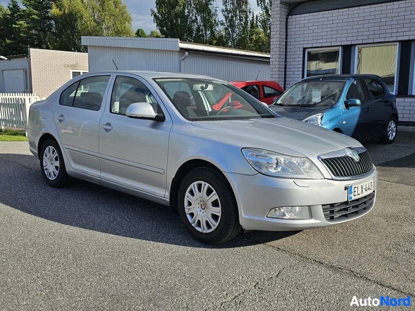 Etuviisto, nokka vasemmalle: Hopeanvärinen nelioven sedan sivulta kuvattu, pysäköity parkkipaikalle. Tummansininen sadeauto ja punainen auto näkyvät taustalla.