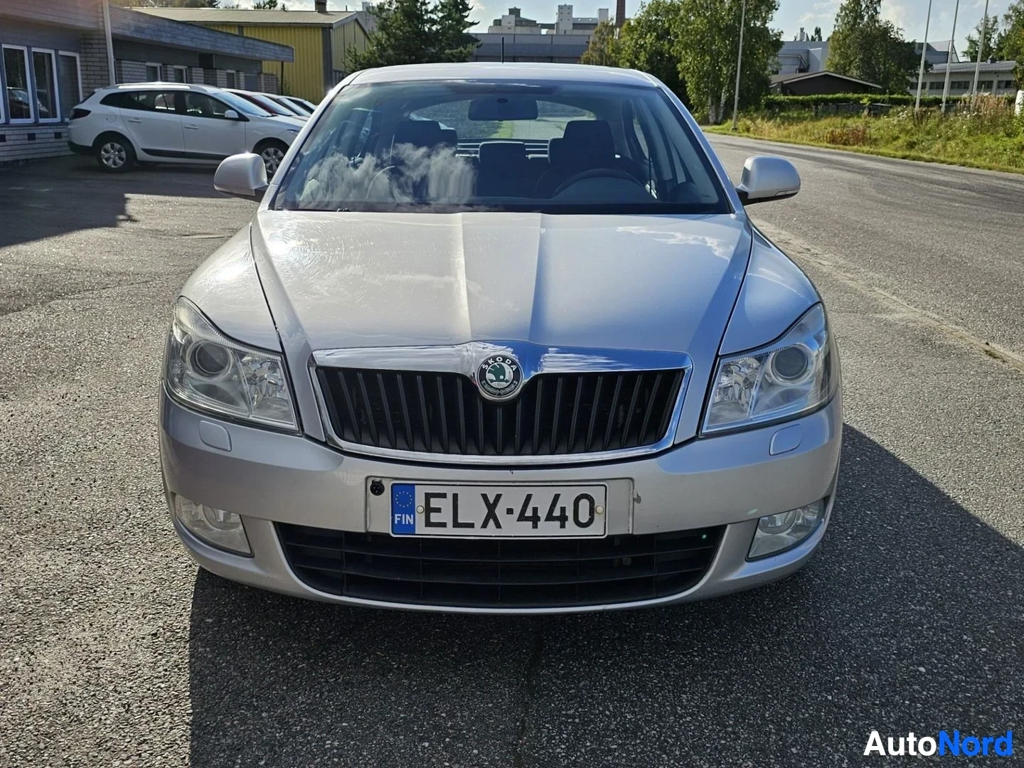 Edestä: Hopeanvärinen nelioven sedan edestä kuvattu parkkipaikalla. Skoda-tunnus näkyvät etuosassa. FIN-tunnus ja rekisterikilvessä