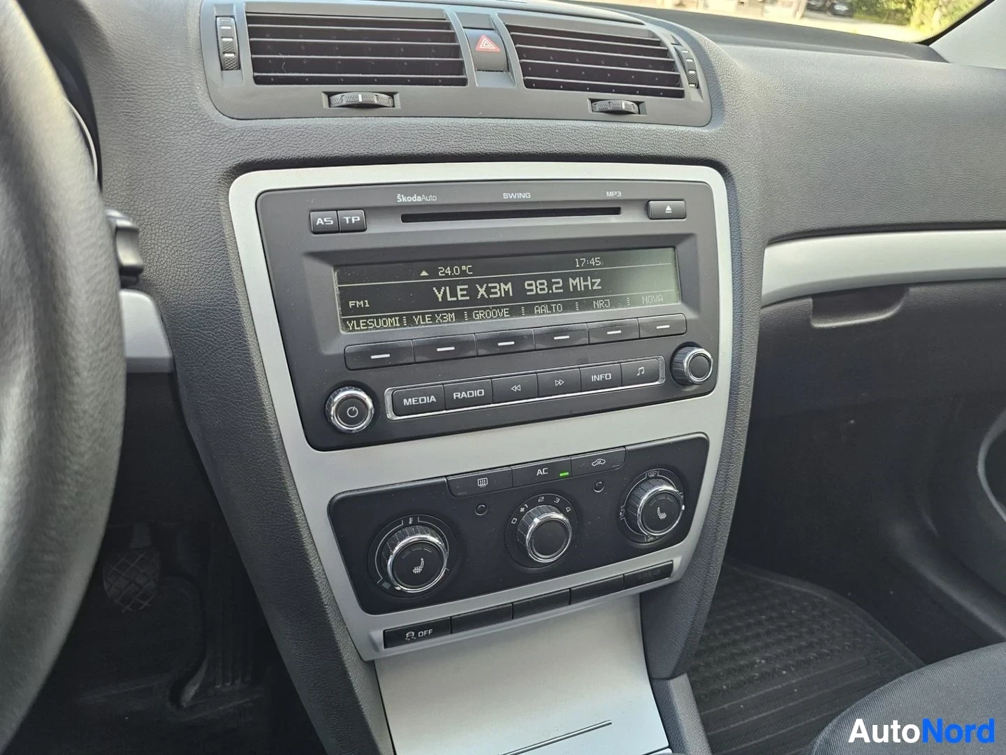 Ohjaamo: Skoda-merkkisen auton sisätilaa, kuva keskikonsolista ja ilmastointi-ohjaimista. Näkyy radio-cd-soitin, ilmanvaihtokontrollit ja lämpötilan säätöpainikkeet.