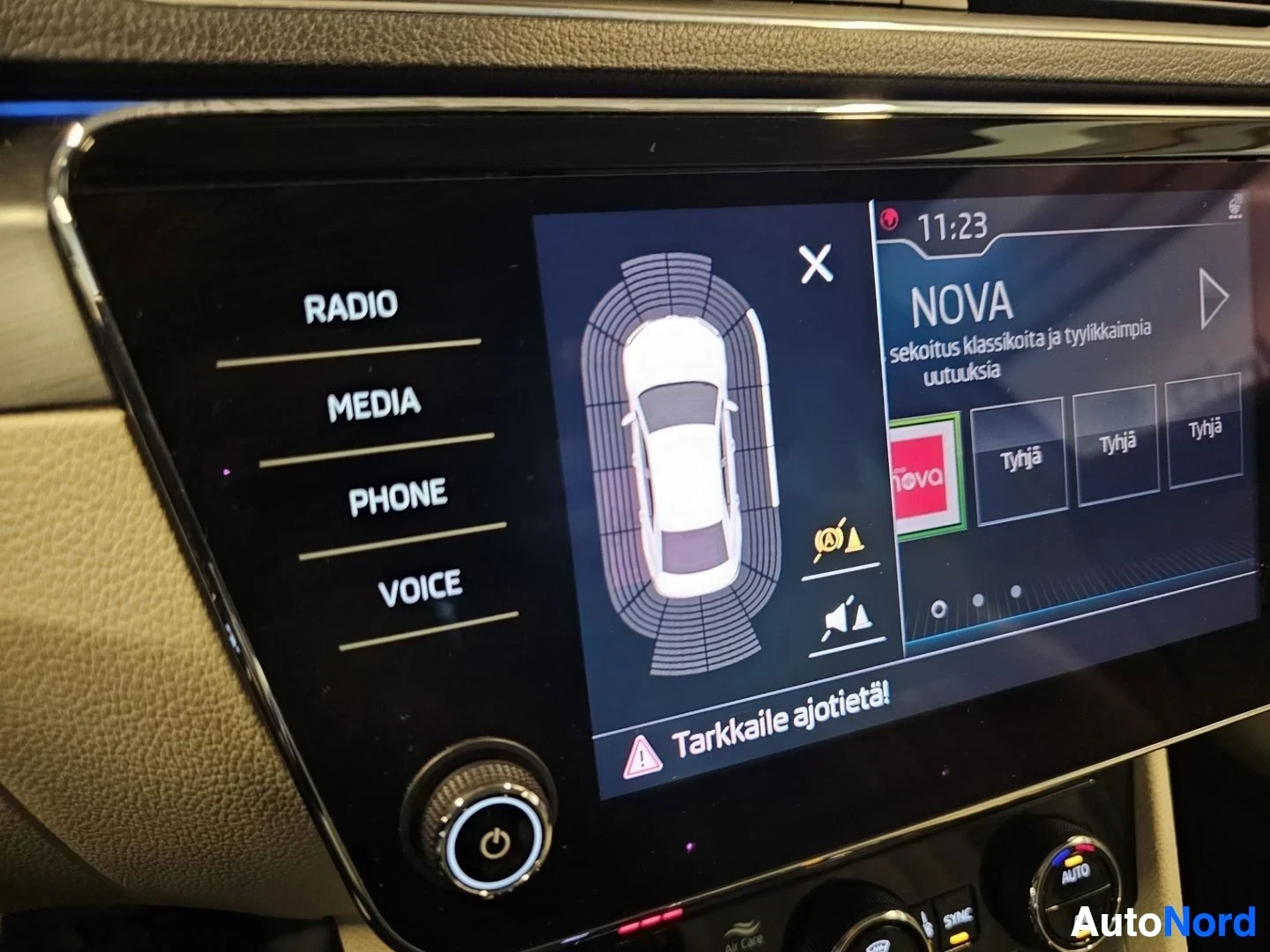 Yksityiskohta: Infotainment-näyttö näyttää radiovalikkoa ja automaattisen paikoituksen kalibrointiruutua NOVA-radiokanavalla; näytön vasemmalla puolella näkyy valikko (Radio,