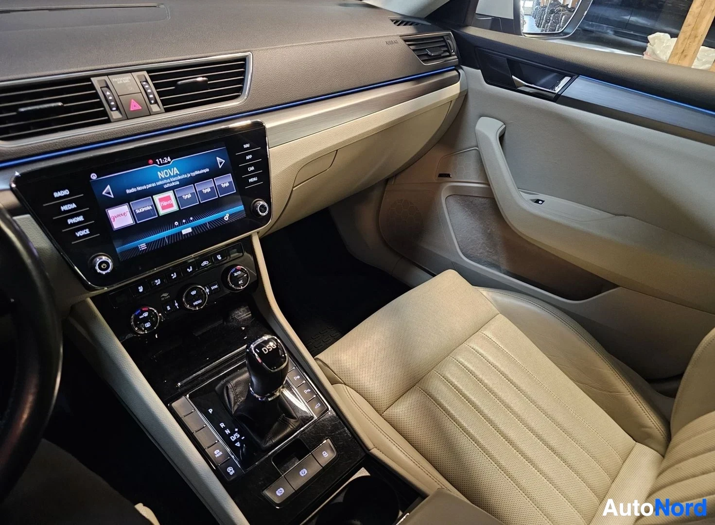 Ohjaamo: Auton sisustus: beige nahkapenkit, infotainment-näyttö, ilmastointi-säädöt ja vaihdelevy käyttäjän näkökulmasta.