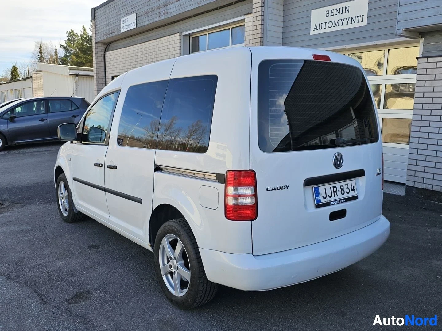 Takaviisto: Valkoinen Volkswagen Caddy näyttää takaa ja vasemmalta puolelta, rekisterikilvessä, taustalla rakennus jonka kyljessä teksti 'BENIN AUTOMAYYNNTI'