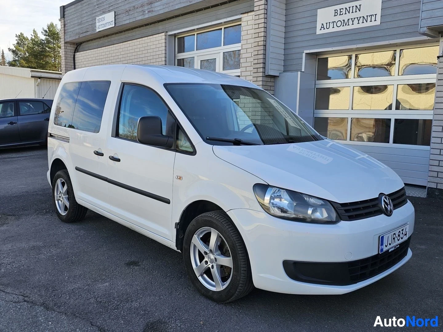 Etuviisto, nokka vasemmalle: Valkoinen Volkswagen Caddy pakkuutiauto kolmen neljänneksen näkymässä vasemmalta, pysäköitynä asfaltoitavalle alueelle toimistotalon edessä.