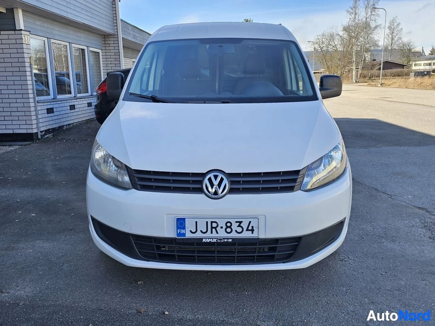 Edestä: Valkoinen Volkswagen Caddy edestä katsottuna, pysäköitynä asfaltoitavalle alueelle rakennuksen viereen.