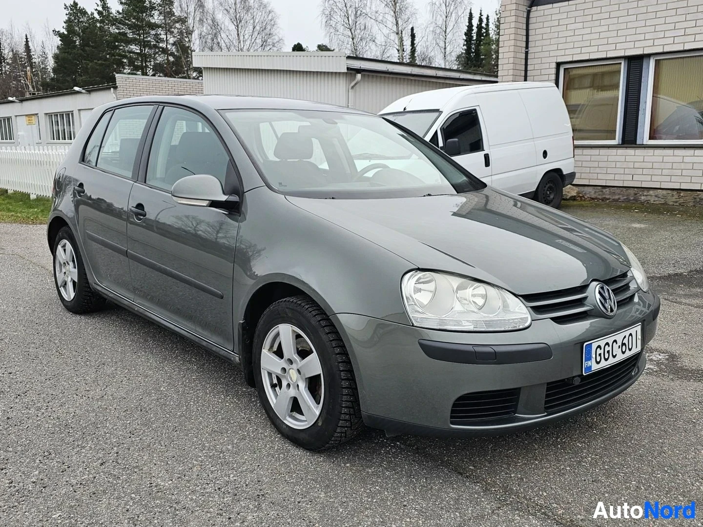 Etuviisto, nokka vasemmalle: Harmaa Volkswagen Golf viistoperä, kuvattu vasemmalta etukolmesta, rekisterikilvessä, valkoisia ketjurenkaita, pysäköity betonialustalle