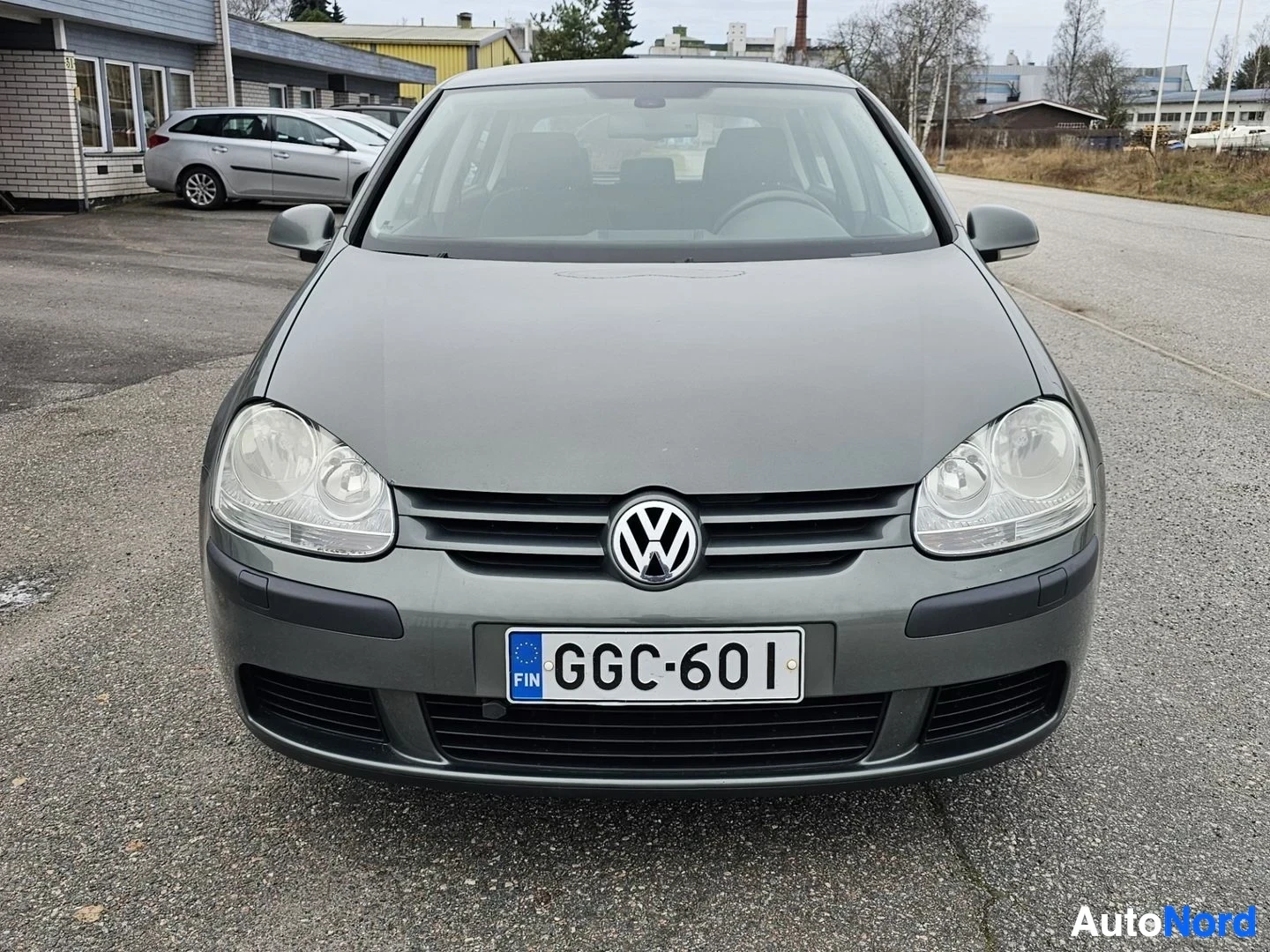 Edestä: Harmaa Volkswagen Golf etunäkymä, rekisterikilvessä, Suomen liput kilven vasemmalla puolella, hallissa pysäköityä valkoista pakettiautoa taustalla