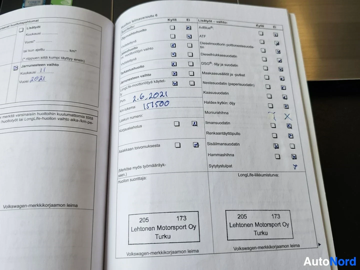 Asiakirja: Volkswagen-huoltokirja avautuneena, sivulla näkyy huollon pvm-merkinnät ja laitteet-tarkistuslista. Kynällä kirjoitettuja päivämääriä 2.6.2021 ja 165100.