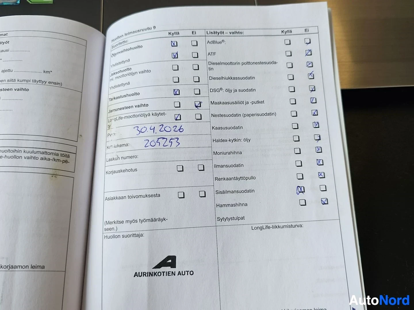 Asiakirja: Volkswagen-huoltokirjan sivu avautuneena, näkyy huoltohistoriaa ja Aurinkoinen Auto -merkkilogo. Dokumentissa merkintöjä kynällä.