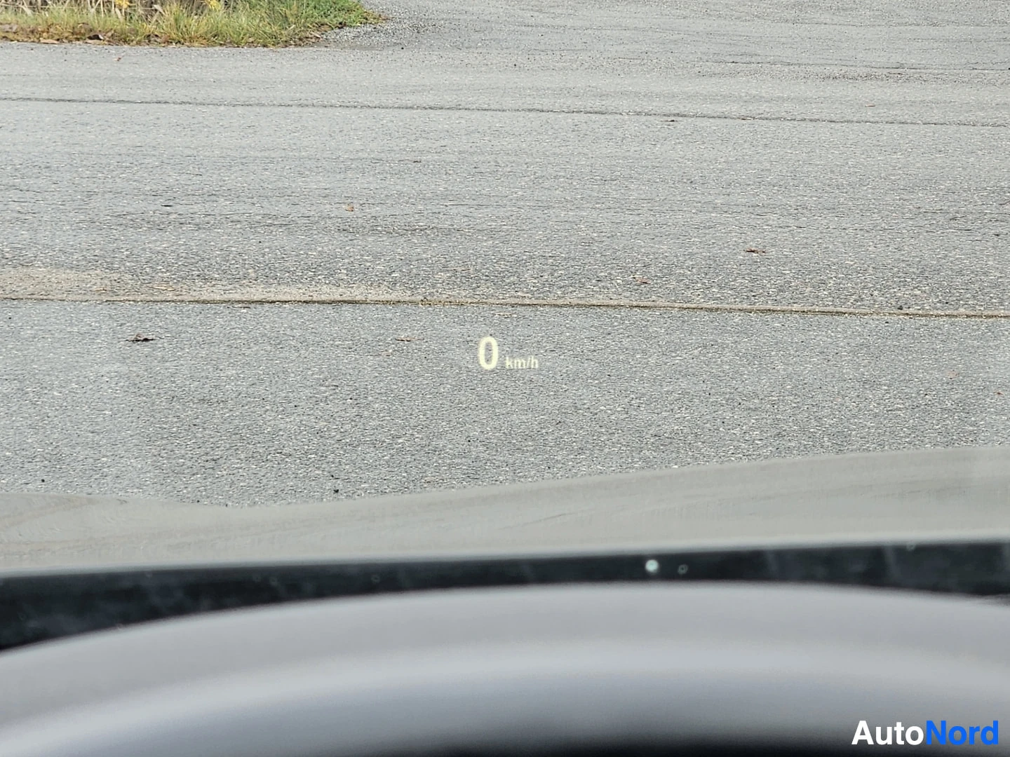 Muu: Auton etupuoli pysäköintialueella, nopeusmittari näyttää 0 km/h, näkyvissä on auton etuosa ja kova asfaltti.