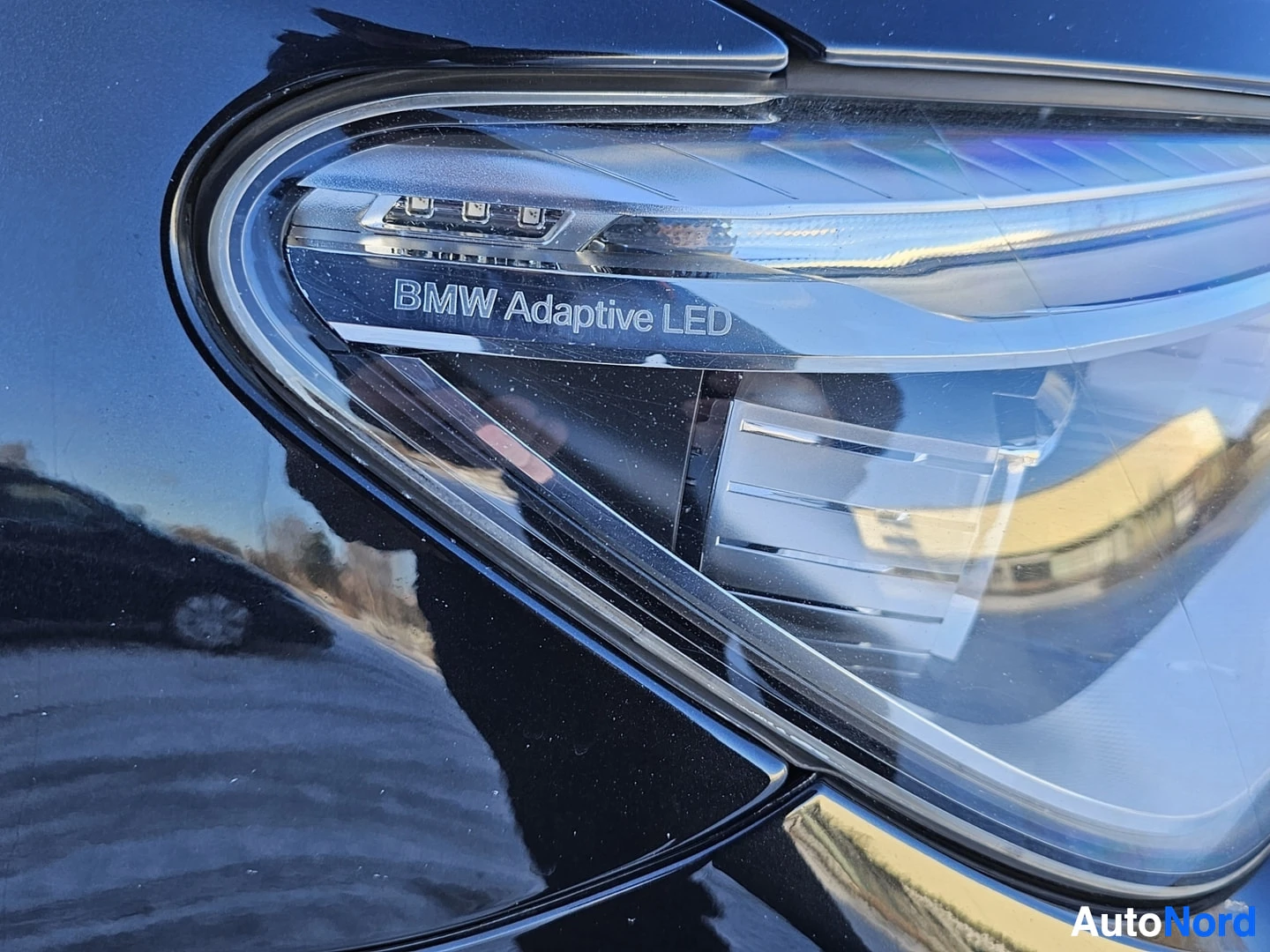 Yksityiskohta: Lähikuva BMW:n vasemmasta ajovalosta, johon on kirjoitettu 'BMW Adaptive LED'. Valotesla näkyy sinisellä taustalla ja pientä heijastusta näkyy.