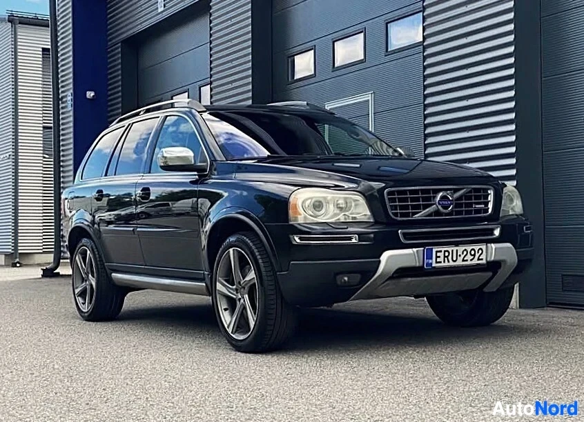 Etuviisto, nokka oikealle: Musta Volvo XC90 kolmikulmainen näkymä vasemmalta puolelta, valkoinen espanjalaislogo näkyy, keskenään valmiit valokiilat ja ammunneet renkaat.