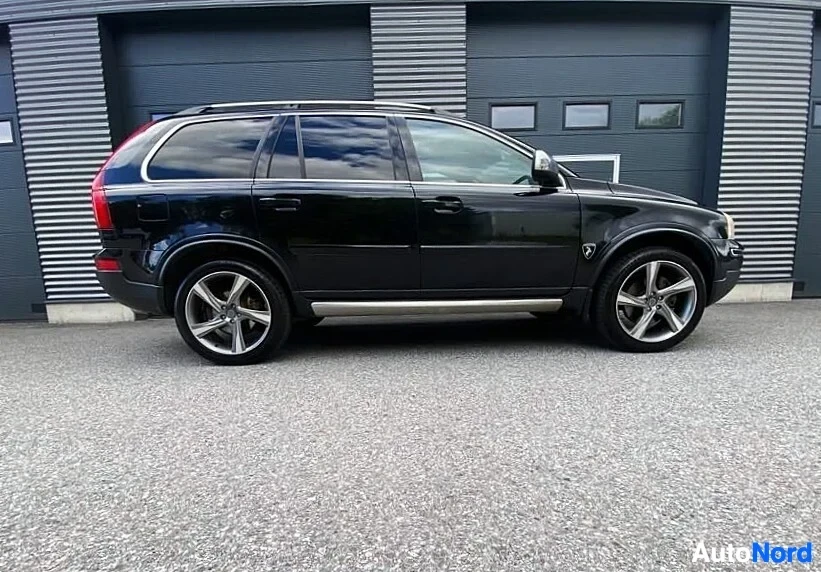 Sivulta: Musta Volvo XC90 -maastoauto sivusta katsottuna, kokonaisuus näkyy, hopeaiset vanteet, metallisen harmaan seinustan edessä