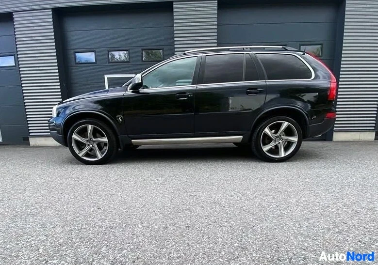 Sivulta: Musta Volvo XC90 -maastoauto sivusta katsottuna, kokonaisuus näkyy, hopeaiset vanteet, metallisen harmaan seinustan edessä