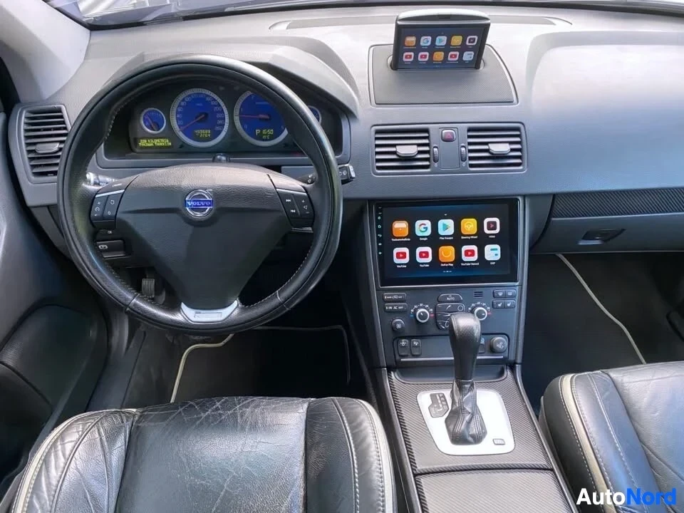 Ohjaamo: Auton sisustus edestä kuvattuna. Näkyy Volvo-logo ohjauspyörässä, digitaaliset mittarit sinisillä näytöillä, päälle asennettu Android-näyttö, ilmastointi-ilmave
