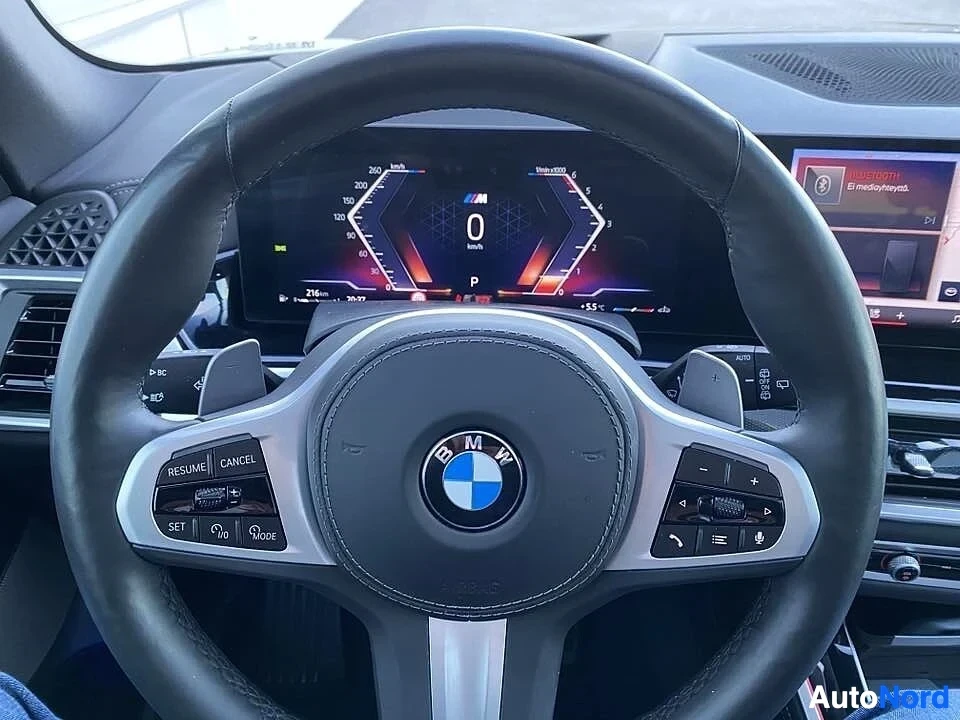 Yksityiskohta: Lähikuva ohjauspyörästä. Näkyy BMW-merkki keskellä, valkoiset M-aksentit, vasemmalla RESUME, CANCEL, SET, Mode-painikkeet, oikealla ääneen ja puhelujen hallinta