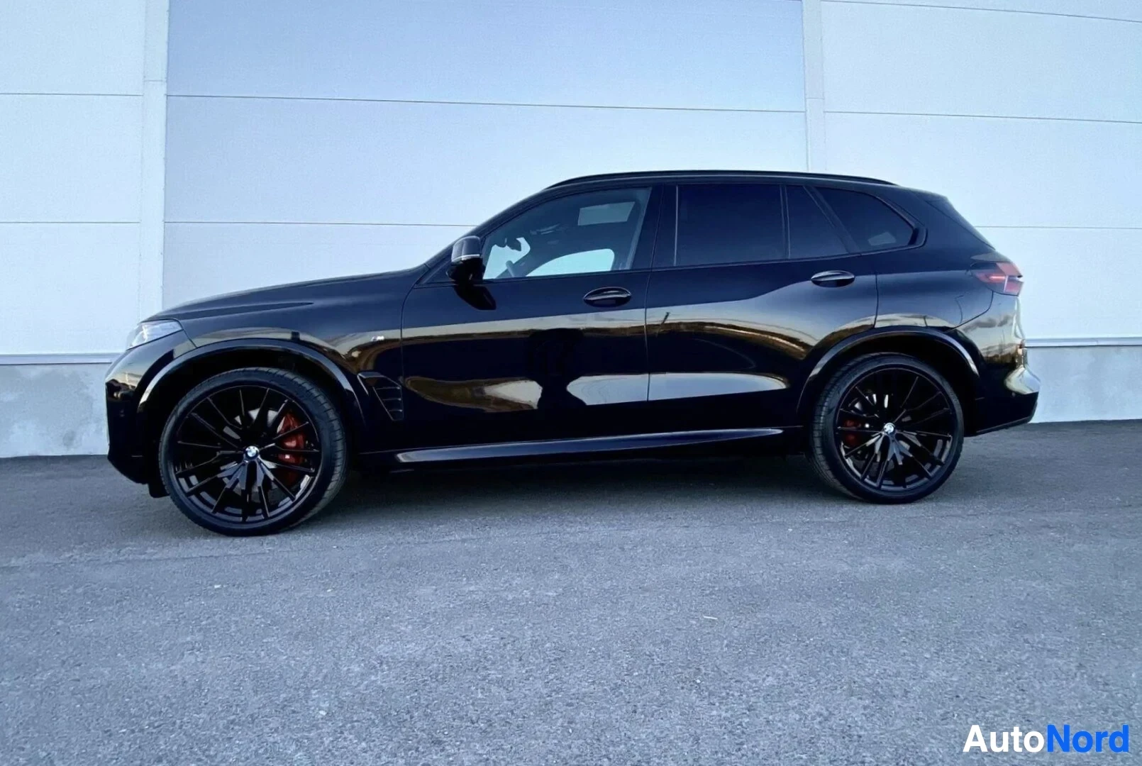 Sivulta: Musta BMW X5 sivusta kuvattuna, näkyy koko ajoneuvo pysäköitynä vaalealla betoniseinustalla