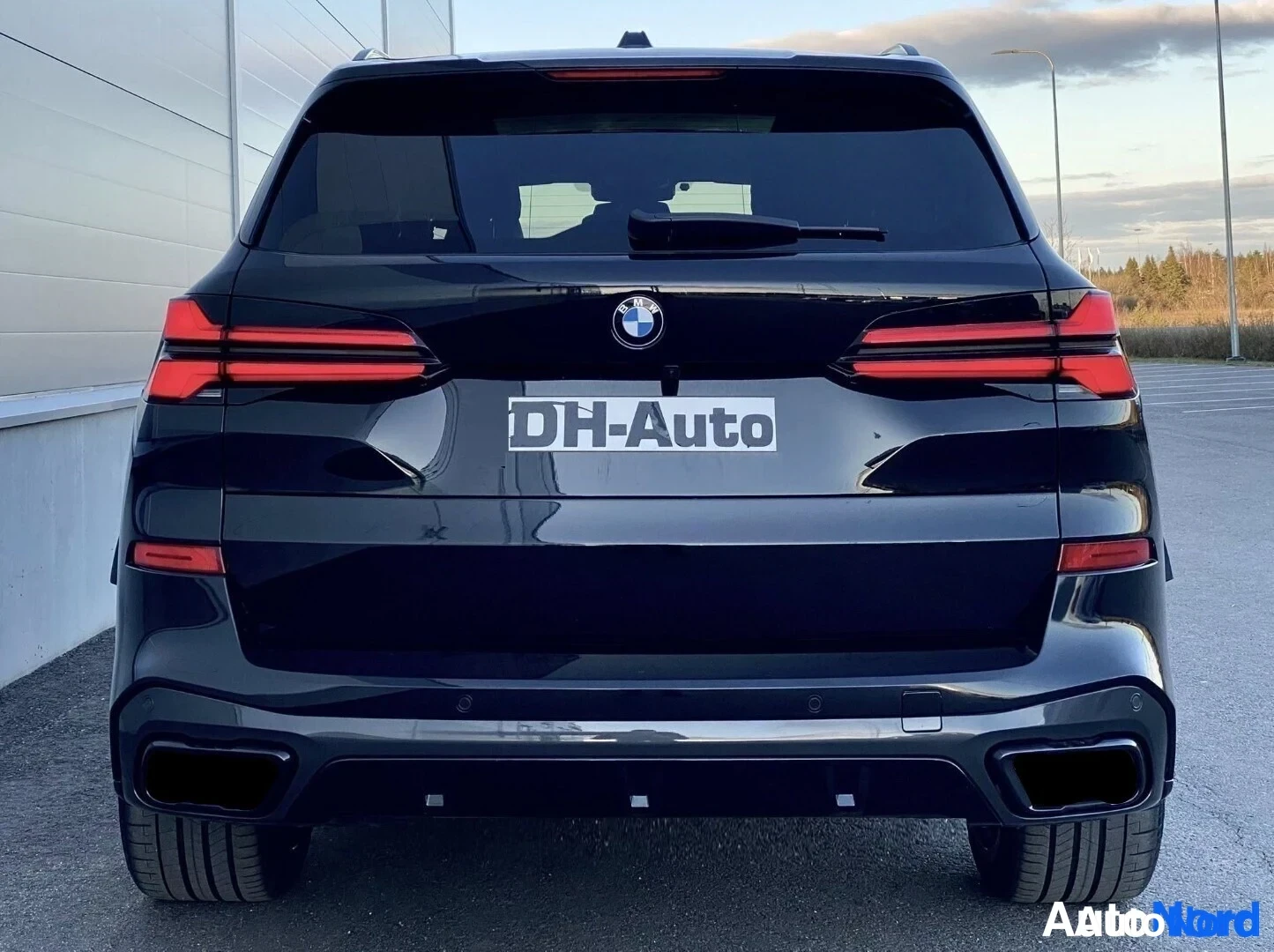 Takaa: Musta BMW X5 -henkilöauto takaa kuvattuna, takavalot päällä. Takaluukun päällä on DH-Auto-kilpi ja alhaalla kaksi pakoputken ulostuloa.