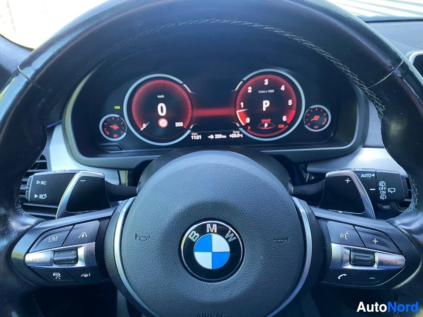 Ohjaamo: BMW:n ohjauspyörä ja digitaalinen mittaristo punaisen valaistuksen kanssa; näytössä näkyy vaihteistoasento P ja nopeusmittari.