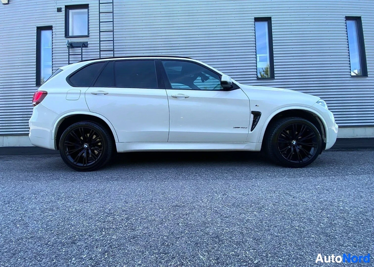 Sivulta: Valkoinen BMW X5 F15 -mallin katumaisen talon seinää vasten, näkyy koko oikea sivu mustan-ja-hopeaksen rengaskierroksin.